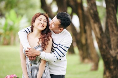 Mutlu Vietnamlı genç adam kız arkadaşına arkadan sarılıyor ve onu yanağından öpüyor.