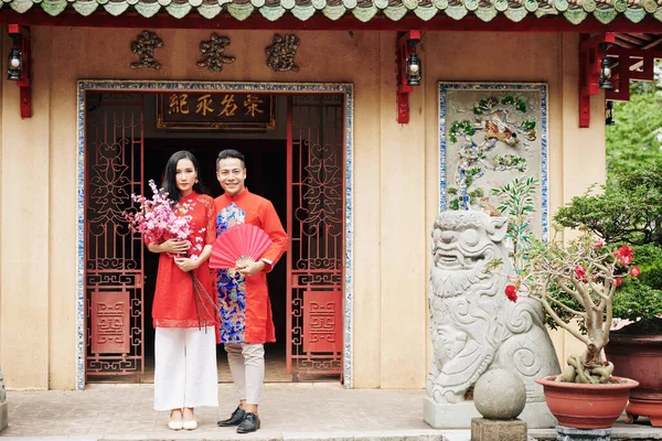 身穿传统服装 快乐的越南年轻夫妇站在寺庙门口 手中拿着盛开的桃枝和纸扇 — 图库照片