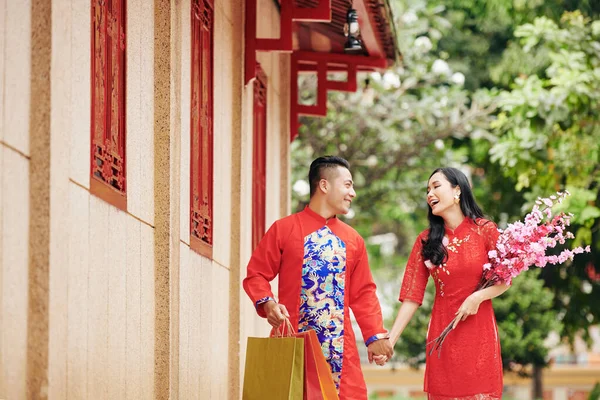 在户外散步时 一对快乐的亚洲年轻夫妇手牵手 身穿传统服装 头戴开花的枝条和购物袋 — 图库照片