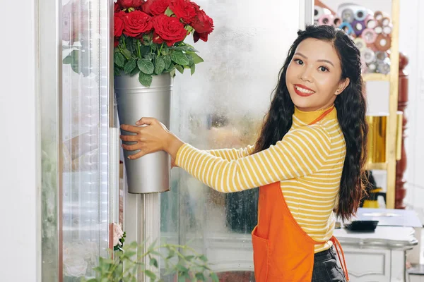 Attractive smiling Asian florist taking bucket of roses out of floral display