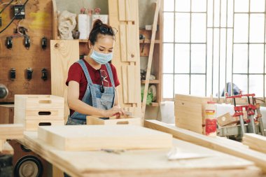 Genç bayan marangoz koruyucu maske takıyor. Çalışma masasında çalışırken tahta blokla parlıyor.