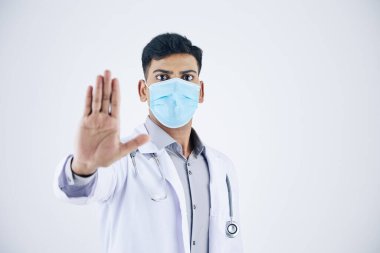 Tıbbi maskeli genç Hintli pratisyen. Durdurma hareketi yapıyor. Beyazlar içinde izole edilmiş.