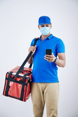 Koruyucu maskeli ciddi teslimatçı mobil uygulamadaki müşteri adresini kontrol ediyor 