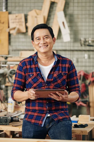 Smiling happy mature carpenter with digital tablet standing at workbench in carpentry workshop