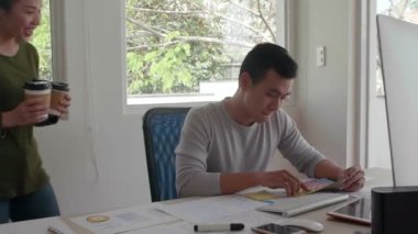 Genç Asyalı adam masaüstünde oturuyor, çalışıyor, sonra kahve bardakları ile bayan meslektaşı geliyor, ona bir tane veriyor, masaya oturuyor ve iletişim kuruyor.