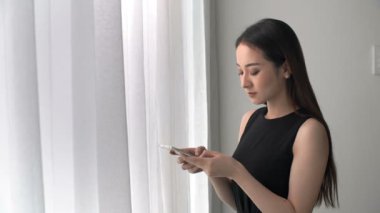 Siyah elbiseli, güzel bir Asyalı kadının elinde akıllı telefonuyla pencerede durup mesaj yazarken orta boy fotoğrafı.