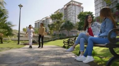 Geniş açıda iki Asyalı genç adamın park boyunca ilerlemesi, iki kızın tahta bankta oturması ve onlara beşlik çakması.