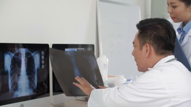 Közepes Felvételű Ázsiai Férfi Orvos Asztalnál Összehasonlítja Röntgenképet Számítógép Képernyőjén — Stock videók