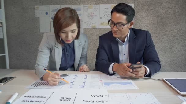 アジアのビジネスマンとビジネスマンの女性が一緒に机の上に座って それの上に横たわるさまざまなチャートや設計図を議論します — ストック動画
