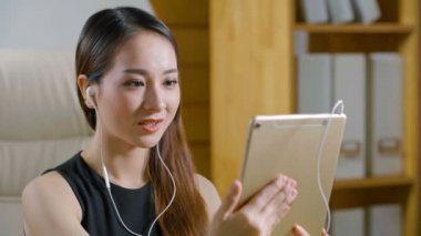 Elinde tabletle oturan ve kulaklıkla video konuşması yapan Asyalı genç bir kadın.