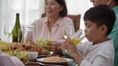 Asyalı bir çocuğun ön planda portakal suyu içip gülümsemesi ve ailesinin arka planda konuşması.