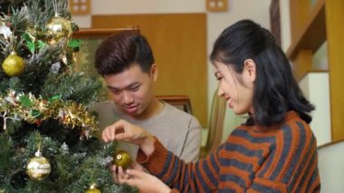 Genç ve çekici Asyalı kadın ve erkek arkadaşı evde Noel ağacını güzel süslerle süsler, konuşur ve neşeyle gülümserler.