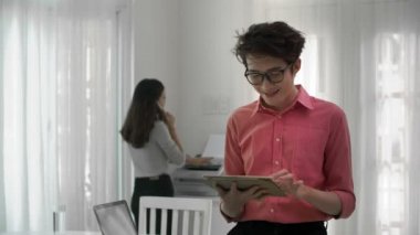 Orta boy Asyalı bir adam elinde tabletle dikilirken, sonra kameraya bakıp gülümserken, arka plan baskısı ve telefon konuşması yaparken.