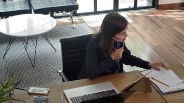 Yüksek açılı iş kadını çalışma masasında otururken, video görüşmesi için kulaklık kullanırken, tablet ve kağıtlardaki bazı bilgileri karşılaştırırken, her şeyi bir kenara koyup dizüstü bilgisayarla yazmaya başlarken.