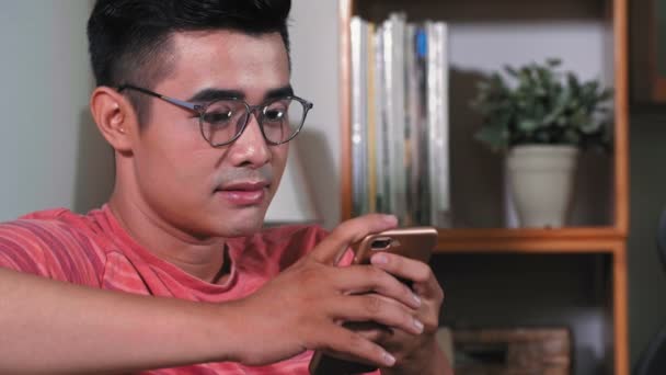 穿着亮晶晶的T恤 戴着眼镜 看着手机里的某个东西 面带微笑的年轻亚洲男人被关了起来 — 图库视频影像