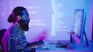 Sakallı genç Hintli programcının bilgisayarda oturup yeni yazılımın kodunu yazarken bilgisayar ekranının ışığı ve kod harfleri ona ve duvarlara yansıyor.