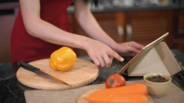 Yemek masasında dikilen, tabletten web sayfalarını araklayan ve sarı dolmalık biberi parçalara ayıran orta boy bir fotoğraf.