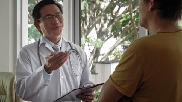 合格的亚洲中年男性医生在检查后与无法辨认的男性病人交谈 让他们放心 并开药方 — 图库视频影像