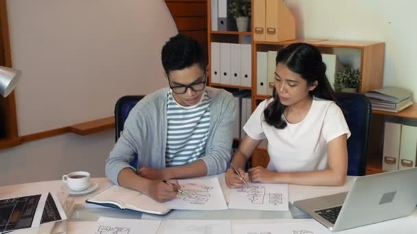若い創造的なアジアの男性はオフィステーブルに座っている間 プロジェクトのスケッチを描き 彼の魅力的な女性同僚にビジネス目標を説明します — ストック動画