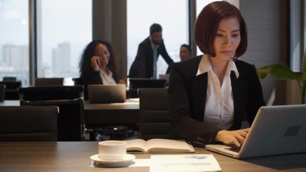 オープンスペースのオフィスで働くスーツを着たアジア人のビジネスマン 彼女はノートパソコンを入力してプランナーにメモを取っています 陽気な女性は電話でチャットやビジネスマンを背景に話して — ストック動画