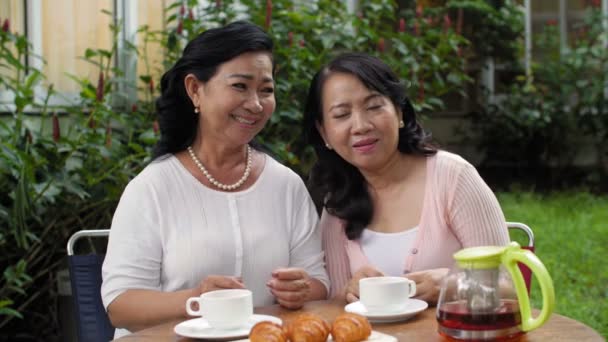迷人的退休亚洲女人与女性朋友一起坐在户外餐桌前 把头靠在肩上 开心地笑着 — 图库视频影像