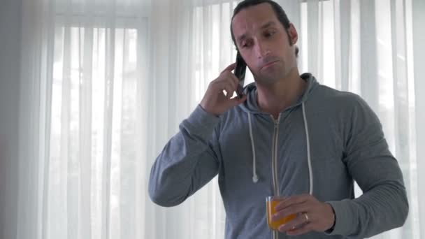 中镜头的白种人站在客厅里 手里拿着一杯橙汁 一边打电话 — 图库视频影像