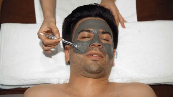 顔の皮膚に粘土マスクを適用するスパベッドと女性の手を保持ブラシに横たわっているアジアの男のトップビュー — ストック動画
