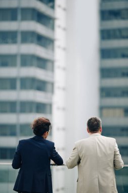 İş arkadaşları çatıda duruyor, şehir manzarasına bakıyor ve şirket gelişimini tartışıyorlar, arkadan bakıyorlar.