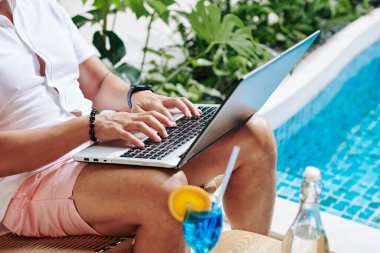 Yüzme havuzunda oturup tatlı kokteyllerin tadını çıkarırken iş adamının laptopta çalıştığı yakın plan görüntüsü.