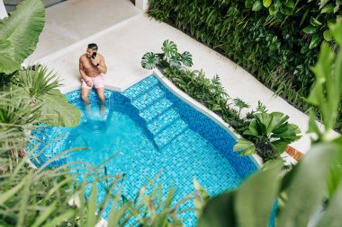 Yüzme havuzunun kenarında oturan yakışıklı bir adam ve telefonla konuşan bir arkadaş ya da iş ortağı, yukarıdan bakan