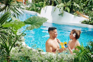 Neşeli Asyalı sevgili ve kız arkadaş yüzme havuzunda duruyorlar yemyeşil bitkiler ve çiçekler arasında, kokteyller içip flört ediyorlar.