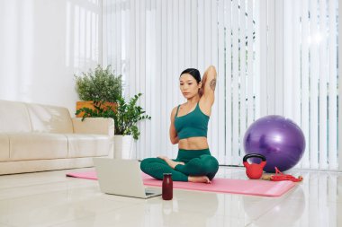 Genç Asyalı kadın dizüstü bilgisayarda video izleyip ellerini arkadan sıkarak inek surat pozu veriyor.