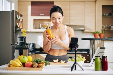 Gülümseyen bir fitness blogcusu mutfak masasında duruyor ve lezzetli sağlıklı kahvaltı smoothie pişirme konusunda online ders veriyor.