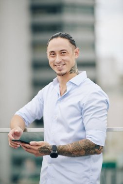 Gömlekli mutlu, melez bir adamın portresi. Arkadaşına ya da meslektaşına mesaj atıyor ve çatıda dikilirken kameraya bakıyor.