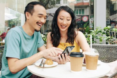 Kafede otururken akıllı telefondan komik mimler ve videolar izleyen mutlu genç Çinli çift.