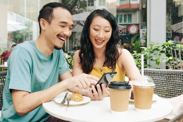 Kafede otururken akıllı telefondan komik mimler ve videolar izleyen mutlu genç Çinli çift.