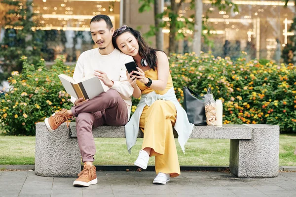 Handsome young Chinese man sitting on bench and reading book when girlfriend leaning on shoulder and texting friends or checking social media