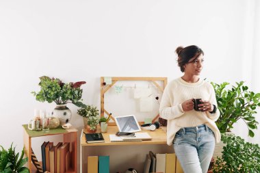 Genç zarif bayan grafik tasarımcısının portresi masasında elinde bir fincan kahveyle duruyor ve başka tarafa bakıyor.