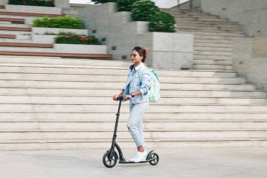 Üniversite kampüsünde elektrikli scooter kullanan, sırt çantalı güzel, şık genç bir kadın, bulanık hareket ediyor.