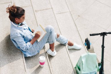 Üniversite kampüsündeki basamaklarda oturan ve eskiz defterinde çizim yapan yetenekli bir bayan grafik tasarım öğrencisi.