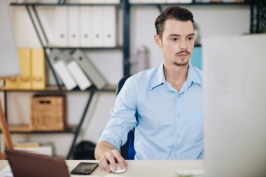 Konsantre genç işadamı ofiste bilgisayar üzerinde çalışıyor ve toplantı için rapor hazırlıyor.