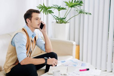 Ciddi genç işadamı, çizelgeler ve raporlarla dolu masada oturuyor ve evden çalışırken iş arkadaşıyla telefonda konuşuyor.