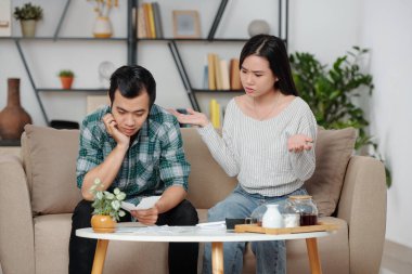Öfkeli genç kadın ve üzgün kocası termanın önünde kahve masasının üzerindeki faturalara bakıyorlar.