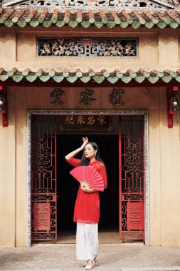 Eski binanın verandasında elinde kırmızı kağıt vantilatörle duran zarif, güzel Asyalı kadın.