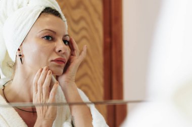 Kafasında havlu olan güzel bir kadın banyoda aynanın önünde duruyor ve nemlendirici losyon sürüyor.