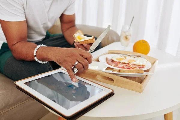Close-up image of man eating breakfast and checking stock market data on screen of tablet computer