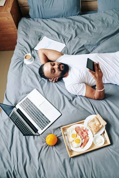 怠惰や疲れた髭の男は トレイにラップトップと朝食でベッドの上で休んで スマートフォンでニュースやメッセージを読んで — ストック写真