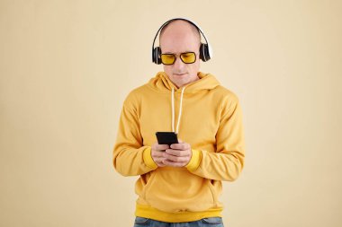 Sarı güneş gözlüklü ve kapüşonlu kel yakışıklı akıllı telefondan müzik dinliyor.