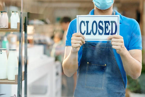 Waiter or shop worker in denim apron standing at entrance and showing closed sign