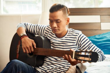 Ciddi genç Vietnamlı adam yurtta gitar çalarak ve şarkı söyleyerek vakit geçiriyor.